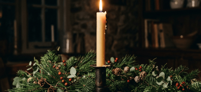 Lit candle in metal holder with pine branches and red berries on wooden table